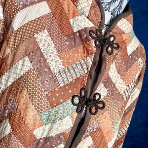 May include: A patchwork jacket in shades of brown, cream, and peach, featuring a geometric pattern. The jacket has a mandarin collar and is fastened with decorative flower-shaped toggles. The fabric appears quilted, adding texture and warmth to the garment.