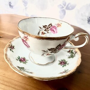 May include: A vintage teacup and saucer set. The white porcelain cup and saucer feature a floral design with pink and brown roses and green leaves. Gold trim accents the edges of the cup and saucer.