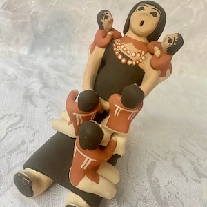 May include: A hand-painted ceramic figurine of a Native American woman with four children. The woman is wearing a black dress with a beaded necklace and has her arms around two children. The other two children are sitting on her lap. The figurine is painted in shades of brown, red, and white.