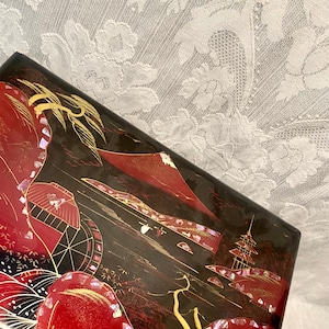 May include: A red and black lacquered wooden box with a Japanese scene painted on the lid. The scene features a mountain, a bridge, and a boat. The box is decorated with gold and mother-of-pearl accents.