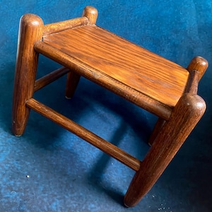 May include: A small, wooden step stool with a rectangular top and four angled legs. The stool is made of a dark-stained wood, showcasing the natural grain. The legs are rounded at the top and bottom, adding a touch of elegance to the design.