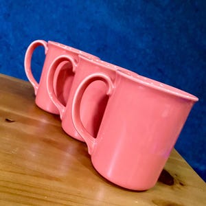 May include: Three coral-coloured ceramic mugs with white polka dots along the rim are arranged on a wooden surface. Each mug has a curved handle. The background is a textured blue surface.
