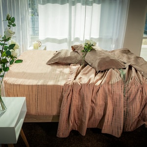 May include: A bedroom scene featuring a bed with a peach-colored striped bedspread and matching pillows. A white side table holds a vase of white roses. Sheer white curtains frame a window, and the room is bathed in natural light.
