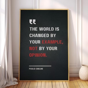May include: Black and white motivational quote poster with gold frame. The quote reads "The world is changed by your example, not by your opinion."  The quote is attributed to Paulo Coelho.