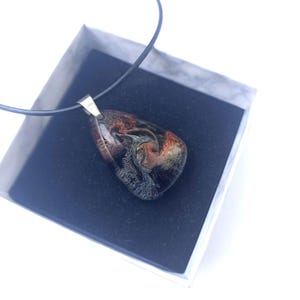 May include: A teardrop-shaped pendant necklace with a black, orange, and silver resin design. The pendant is hanging from a black cord and is in a white box with a black interior.
