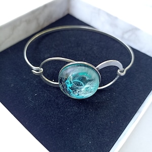 May include: A silver bangle bracelet with a round, turquoise blue glass cabochon featuring a white wave design.
