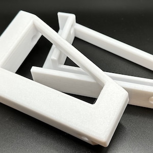 Puede incluir: Dos soportes de plástico blanco impresos en 3D con una abertura rectangular. Los soportes están diseñados para montarse en una pared u otra superficie.
