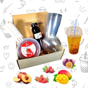 Puede incluir: Una caja de regalo llena de ingredientes para hacer bubble tea. La caja contiene una botella de jarabe de té negro, un recipiente de perlas de tapioca, una taza con tapa y pajitas. La caja está rodeada de fruta, incluyendo mango, maracuyá, melocotones, frambuesas y fresas.