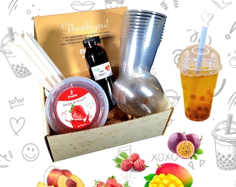 Op de afbeelding: Een cadeaudoos gevuld met ingrediënten om bubble tea te maken. De doos bevat een fles zwarte theesiroop, een container met tapiocaparels, een beker met deksel en rietjes. De doos is omringd door fruit, waaronder mango, passievrucht, perziken, frambozen en aardbeien.