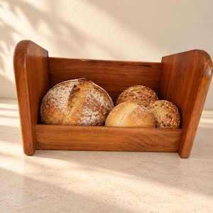 May include: A rectangular wooden bread bin with high side panels containing one large round loaf of crusty bread and three smaller round bread rolls. The wooden container features a warm brown tone and a smooth finish, holding the baked goods.