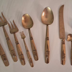 May include: A collection of antique silverware with a gold finish. The set includes forks, spoons, a knife, and a ladle. The handles have decorative carvings. The silverware is arranged on a light-colored surface.