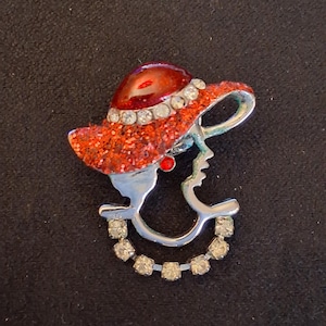 Op de afbeelding: Een vintage broche met een vrouwenprofiel met een rode glitterhoed, versierd met strass-steentjes. De zilverkleurige broche heeft een gebogen ontwerp met meer strass-steentjes. Een kleine rode edelsteen zit bij het oor.