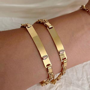 May include: Two gold-colored bracelets with rectangular nameplate designs. Each bracelet features a chain link band and a small oval charm. The bracelets are displayed on a person's arm.