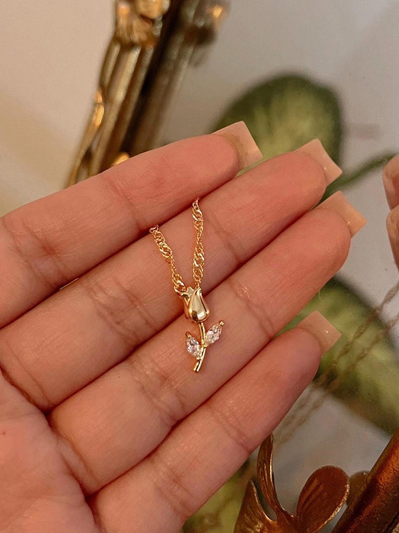 Peut inclure: Un d&eacute;licat collier dor&eacute; avec un pendentif en forme de tulipe. Le pendentif pr&eacute;sente un bouton de tulipe d&eacute;taill&eacute; et deux feuilles orn&eacute;es de petites pierres pr&eacute;cieuses transparentes. La cha&icirc;ne est de conception fine et torsad&eacute;e.