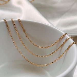 Peut inclure: Trois colliers dor&eacute;s de diff&eacute;rents mod&egrave;les sont pr&eacute;sent&eacute;s dans un bol blanc. Les cha&icirc;nes pr&eacute;sentent divers motifs de maillons, dont un style Figaro et un design torsad&eacute;. Les colliers sont pos&eacute;s sur un fond blanc et doux.