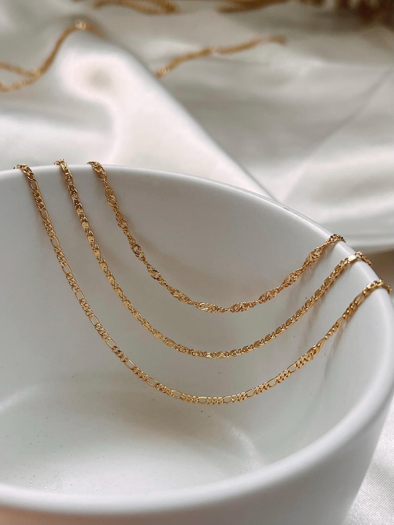 Peut inclure: Trois colliers dor&eacute;s de diff&eacute;rents mod&egrave;les sont pr&eacute;sent&eacute;s dans un bol blanc. Les cha&icirc;nes pr&eacute;sentent divers motifs de maillons, dont un style Figaro et un design torsad&eacute;. Les colliers sont pos&eacute;s sur un fond blanc et doux.