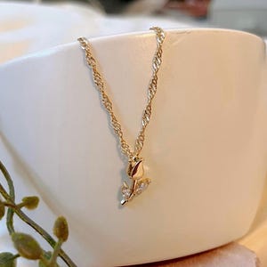 Peut inclure: Un collier dor&eacute; avec une cha&icirc;ne fine et un pendentif en forme de rose. La rose est orn&eacute;e de petites pierres scintillantes. Le collier est pr&eacute;sent&eacute; dans un bol en c&eacute;ramique blanc.