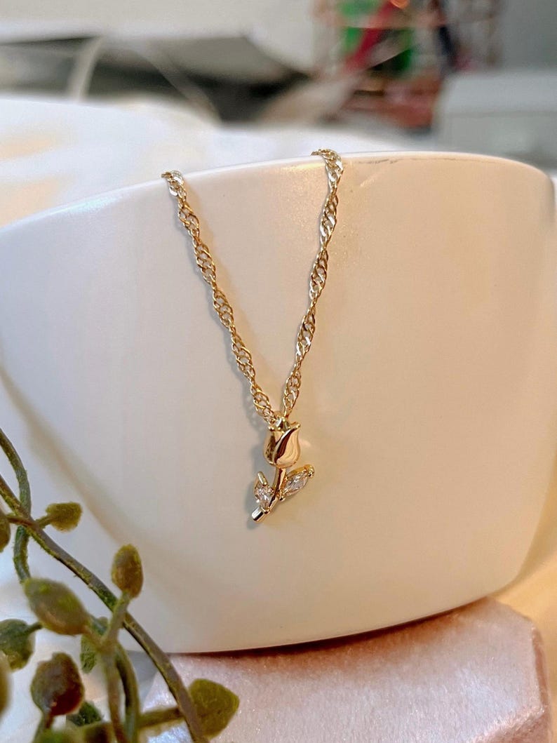 Peut inclure: Un collier dor&eacute; avec une cha&icirc;ne fine et un pendentif en forme de rose. La rose est orn&eacute;e de petites pierres scintillantes. Le collier est pr&eacute;sent&eacute; dans un bol en c&eacute;ramique blanc.