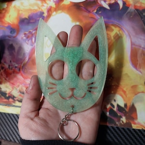May include: A green and white cat-shaped keychain with a silver ring. The keychain is made of a translucent material and has a glitter finish.