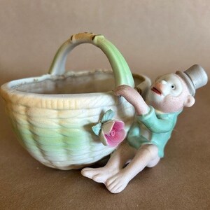 May include: A ceramic planter shaped like a basket with a green and white striped design. A small monkey figurine is holding onto the basket handle. The monkey is wearing a green shirt and a black top hat. There is a pink flower on the side of the basket.