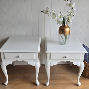 May include: Two white bedside tables with a single drawer and cabriole legs. The tables are on a wood floor and a vase with white flowers is in the background.