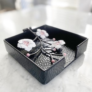 May include: Black napkin holder with a hammered finish and a metal branch design with pink and white flowers.
