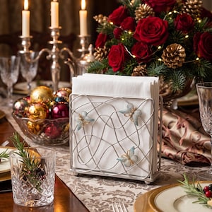 May include: A silver-toned metal napkin holder featuring a wire-frame design with three decorative butterflies attached to the front. The holder contains a stack of plain white square paper napkins, standing upright on a flat rectangular base.