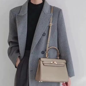 May include: A light gray leather handbag with a gold clasp and a long strap. The bag is being held by a person wearing a gray coat.