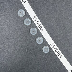 May include: Five clear, round, silicone bottle stoppers on a gray surface. A white ribbon with the word "SATTIARA" printed on it is diagonally across the image.
