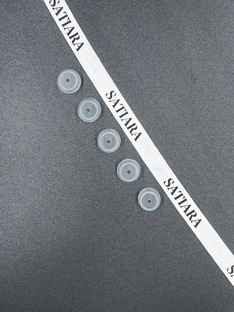 May include: Five clear, round, silicone bottle stoppers on a gray surface. A white ribbon with the word "SATTIARA" printed on it is diagonally across the image.