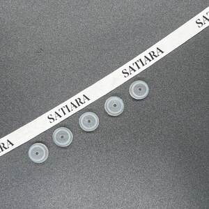 May include: Five white rubber bottle stoppers with a black ring around the center. The stoppers are arranged in a row on a gray surface. A white ribbon with the text "SATIARA" is visible in the background.