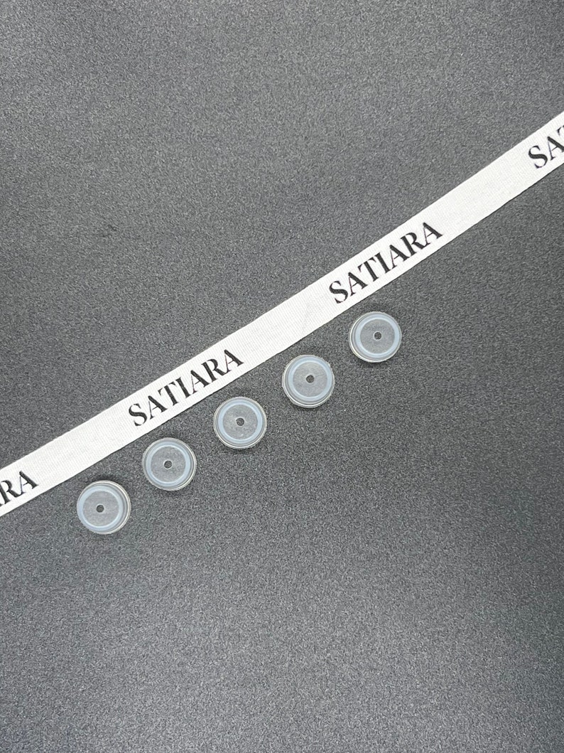 May include: Five white rubber bottle stoppers with a black ring around the center. The stoppers are arranged in a row on a gray surface. A white ribbon with the text "SATIARA" is visible in the background.