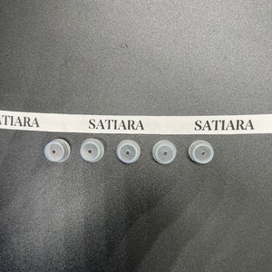 May include: Five small, round, gray rubber stoppers with a small hole in the center. The stoppers are arranged in a row on a black surface. A white ribbon with the text "SATIARA" printed on it is above the stoppers.