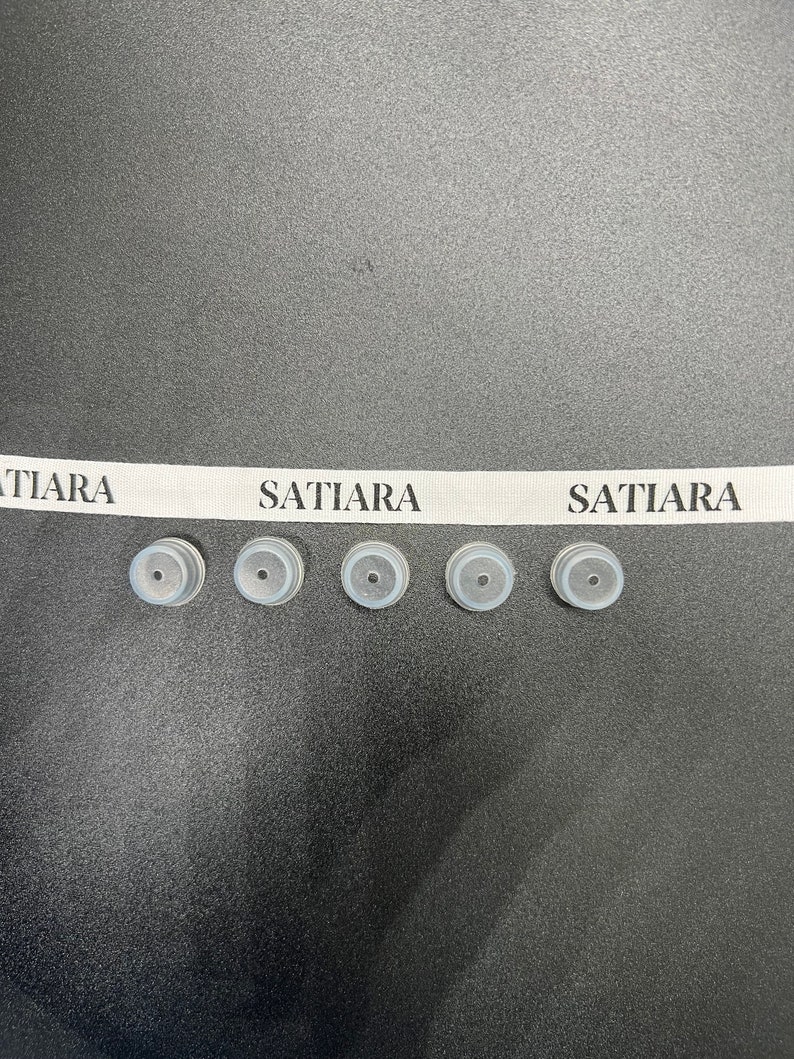 May include: Five small, round, gray rubber stoppers with a small hole in the center. The stoppers are arranged in a row on a black surface. A white ribbon with the text "SATIARA" printed on it is above the stoppers.