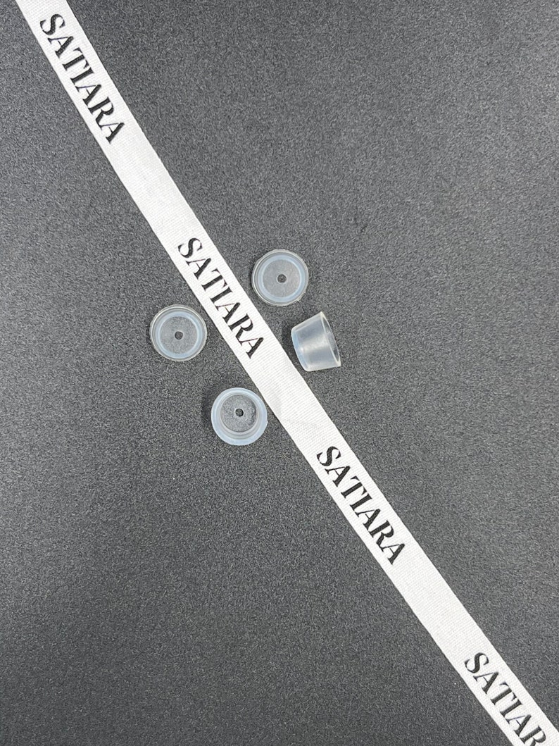 May include: Four clear plastic bottle stoppers on a gray surface with a white ribbon that says "SATIARA" on it.