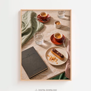 May include: A framed print showing a still life arrangement with coffee cups, saucers, and an eclair on a plate. A book titled "The Metamorphosis" by Franz Kafka is also present. The scene is set on a marble-effect surface with a green cloth.