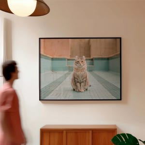 May include: A framed photograph of a cat sitting in a turquoise-tiled pool. The cat has long, light brown fur and upright ears. The photograph is displayed on a white wall above a wooden cabinet.