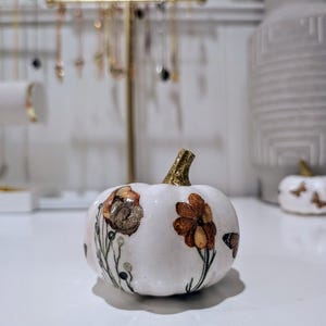 Puede incluir: Una pequeña calabaza de cerámica blanca con diseños florales y de mariposas. La calabaza tiene un tallo dorado con purpurina y está decorada con flores marrones y naranjas y pequeñas mariposas. La calabaza está sobre una superficie blanca.