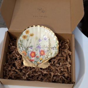 May include: A decorative scallop shell dish featuring a colorful wildflower print on an off-white background with a metallic gold rim and base. The shell is nestled in brown paper crinkle fill inside a cardboard box labeled TILLYPAD.