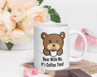 Op de afbeelding: Een witte keramische koffiemok met een bruine cartoonbeer die een knipoog geeft en een kopje koffie vasthoudt. De mok heeft de tekst "Bear With Me, It's Coffee Time!" erop gedrukt.