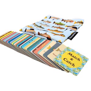 May include: A set of colorful cards with fish illustrations and the text "Match & Catch" on a yellow circle. The cards are fanned out and stacked on a table. A blue and white fabric bag with fish illustrations is in the background.