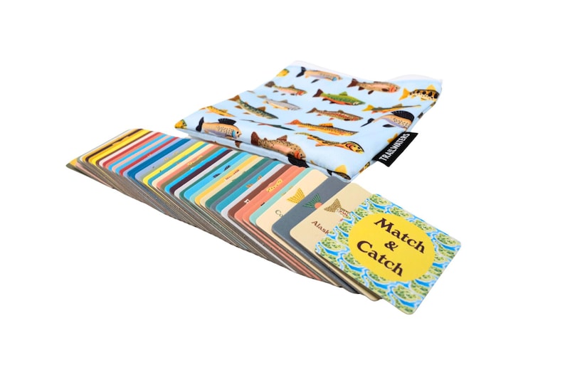 May include: A set of colorful cards with fish illustrations and the text "Match & Catch" on a yellow circle. The cards are fanned out and stacked on a table. A blue and white fabric bag with fish illustrations is in the background.