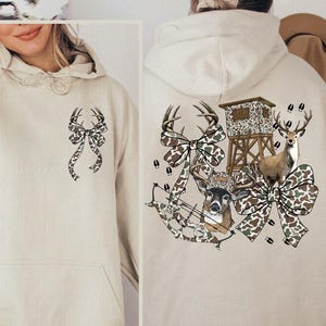 May include: A sand-colored hoodie with a front pocket and a drawstring hood. The front features a bow design with deer antlers. The back of the hoodie displays a hunting-themed graphic with deer, a bow, and a hunting blind.