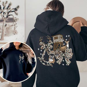 May include: Black hooded sweatshirt with a hunting-themed graphic on the back. The design features a deer, a hunting blind, and camouflage bows. The sweatshirt has a front pocket and a drawstring hood.