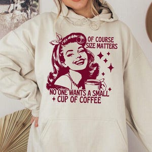 May include: Sand-colored hoodie with a front graphic. The graphic features a retro illustration of a woman with the text "OF COURSE SIZE MATTERS" and "NO ONE WANTS A SMALL + CUP OF COFFEE" in burgundy.