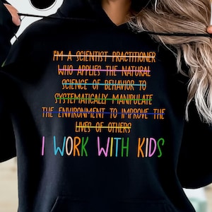 May include: Black hooded sweatshirt with the text "I'M A SCIENTIST PRACTITIONER WHO APPLIES THE NATURAL SCIENCE OF BEHAVIOR TO SYSTEMATICALLY MANIPULATE THE ENVIRONMENT TO IMPROVE THE LIVES OF OTHERS I WORK WITH KIDS" in colorful lettering. The word "BLACK" is in a black circle.