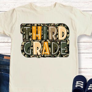 Puede incluir: Una camiseta color crema con las palabras "THIRD GRADE" en un diseño de camuflaje. La camiseta se combina con vaqueros azules, zapatos azul marino y coloridos huevos de Pascua. También es visible el texto "colors can be slightly darker or lighter due to photographing".