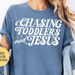 May include: Blue jean-colored t-shirt with the text "CHASING TODDLERS and JESUS" in white. The shirt has rolled-up sleeves and a relaxed fit. A yellow circle with "MORE COLOR OPTIONS" is visible.