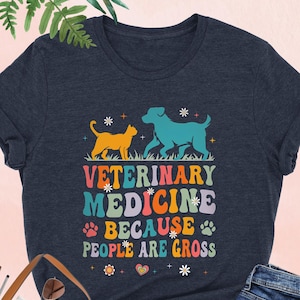 May include: Heather navy t-shirt with a colorful graphic. The graphic features a cat and dog, the words "VETERINARY MEDICINE BECAUSE PEOPLE ARE GROSS", and decorative elements. The shirt is paired with blue jeans and a brown tote bag.