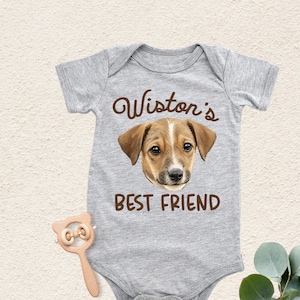 May include: A heather gray short-sleeve baby bodysuit featuring a brown graphic of a dog's face centered between the brown text Wiston's and BEST FRIEND. The garment has a round neckline and snap closures at the bottom for easy access.